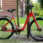Rotes Lite Step 2 Fahrrad mit tiefem Einstieg, Gepäckträger und Schutzblechen, abgestellt im Freien auf grasigem Untergrund neben einem Baum.
