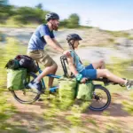 Zwei Personen, Mann und Frau, fahren auf einem blauen Hase Pino Tandem durch eine ländliche, hügelige Landschaft, beladen mit Gepäcktaschen und Schlafsäcken.