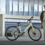 Eine Frau in grüner Kleidung steht neben einem hellgrünen TENWAYS CGO600 Pro E-Bike, das auf einer Betonfläche vor einem modernen Gebäude abgestellt ist.