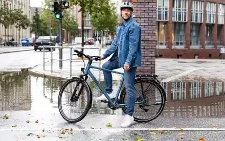Mann mit Helm und Jeans steht neben einem blauen Aviolo 8.1 HT Tourenrad auf nasser Straße mit Spiegelungen.
