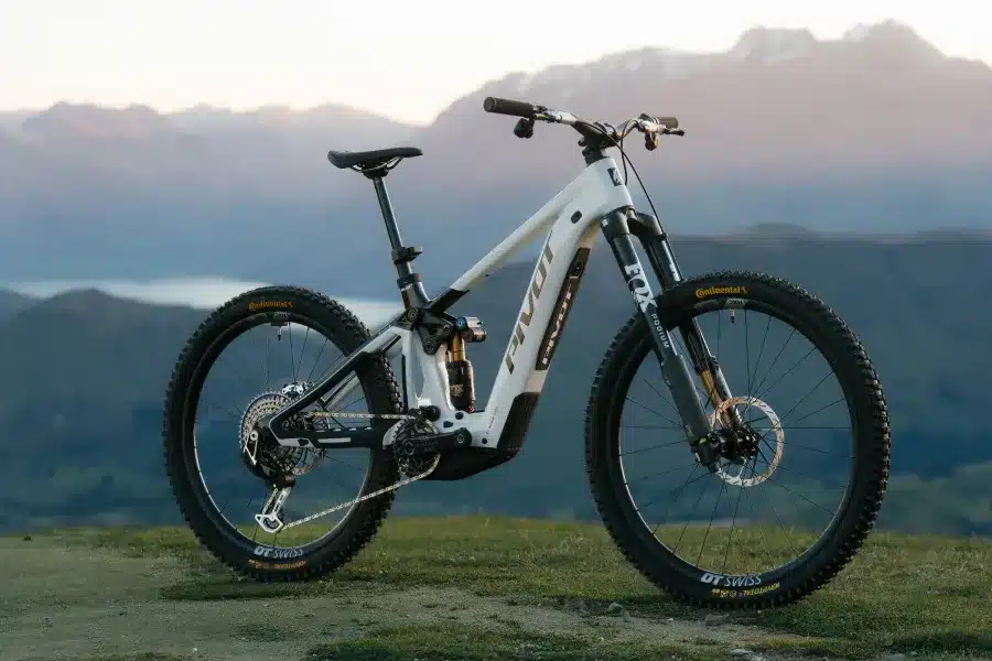 Weißes Pivot Shuttle LT E-Mountainbike steht auf einem felsigen Untergrund mit Bergpanorama im Hintergrund.