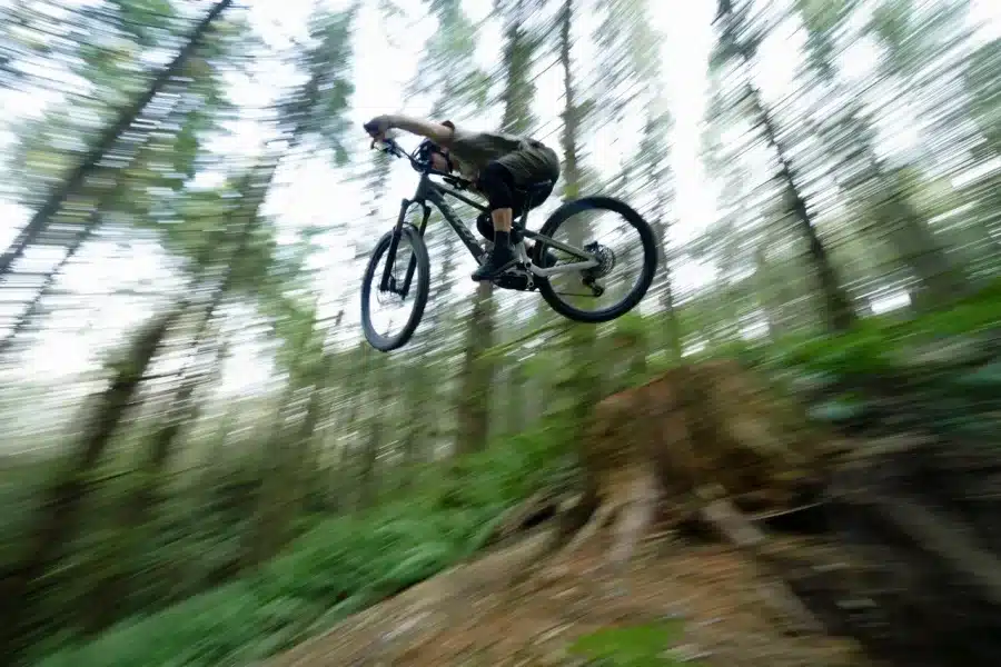 Ein Mountainbiker mit dunklem Trikot und Shorts, der mit seinem Fahrrad in der Luft ist, während er eine Abfahrt in einem Wald fährt.