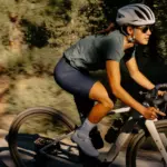 Eine Frau fährt ein Specialized Turbo Creo 2 Expert E-Gravelbike auf einem sonnigen Schotterweg, umgeben von Bäumen und Vegetation.