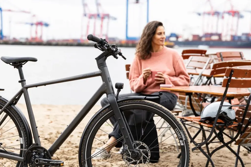 Graues Stevens Caleta 9.1 HT Trekkingrad mit Frau in rosafarbenem Pullover, die an einem Tisch sitzt, im Hintergrund Hafenkranen.