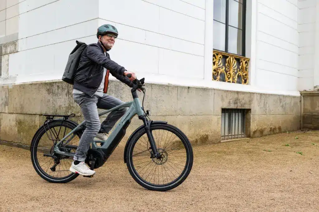 Mann mit Helm und Rucksack fährt ein Stevens E-Triton 7.8.1 Plus Forma E-Bike in River Stone Farbe vor einer Stuckfassade.