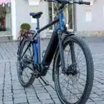 MAXX Crossmaxx ELS E-Trekkingbike, blau, mit Gepäckträger und Seitenständer, steht auf Kopfsteinpflaster.