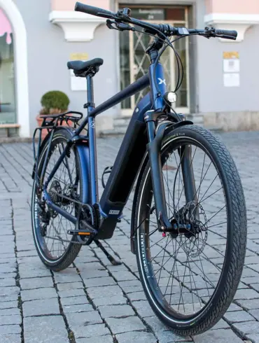 MAXX Crossmaxx ELS E-Trekkingbike, blau, mit Gepäckträger und Seitenständer, steht auf Kopfsteinpflaster.
