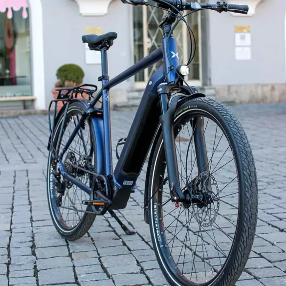 MAXX Crossmaxx ELS E-Trekkingbike, blau, mit Gepäckträger und Seitenständer, steht auf Kopfsteinpflaster.