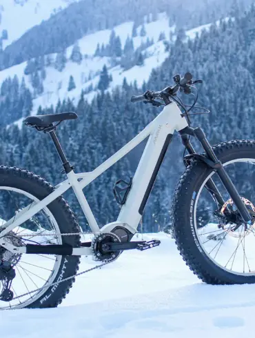 Ein weißes E-Fatbike MAXX Jagamoasta ELS SH steht in tiefer Schneedecke. Im Hintergrund ist eine verschneite Bergkulisse mit Nadelbäumen zu sehen.