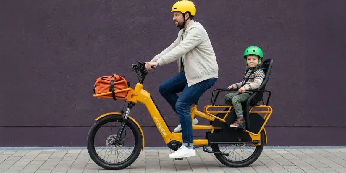 Ein Mann fährt ein gelbes Maxx Maxximo Urban Longtail-E-Bike mit einem Kind, das in einem Kindersitz auf dem Gepäckträger sitzt. Der Mann trägt einen gelben Helm, das Kind trägt einen grünen Helm. Vorne am Rad ist eine orangefarbene Tasche befestigt.