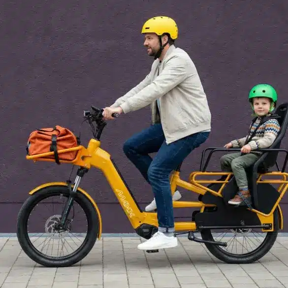 Ein Mann fährt ein gelbes Maxx Maxximo Urban Longtail-E-Bike mit einem Kind, das in einem Kindersitz auf dem Gepäckträger sitzt. Der Mann trägt einen gelben Helm, das Kind trägt einen grünen Helm. Vorne am Rad ist eine orangefarbene Tasche befestigt.