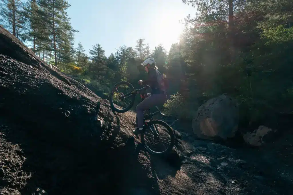 Eine Frau mit Helm und Mountainbike fährt einen felsigen, steilen Hang bergauf, umgeben von Bäumen und natürlicher Landschaft.