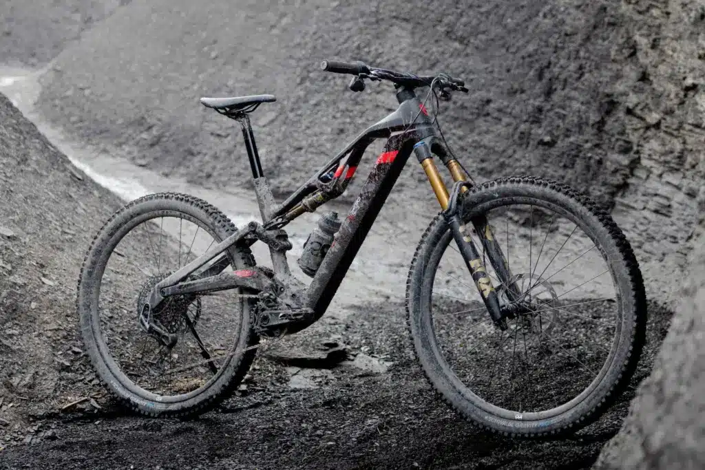 Ein fully-suspension E-Mountainbike der Marke ROTWILD, Modell R.XX, mit Carbonrahmen und integriertem Akku, steht im Gelände. Es ist schlammig und zeigt deutliche Gebrauchsspuren.