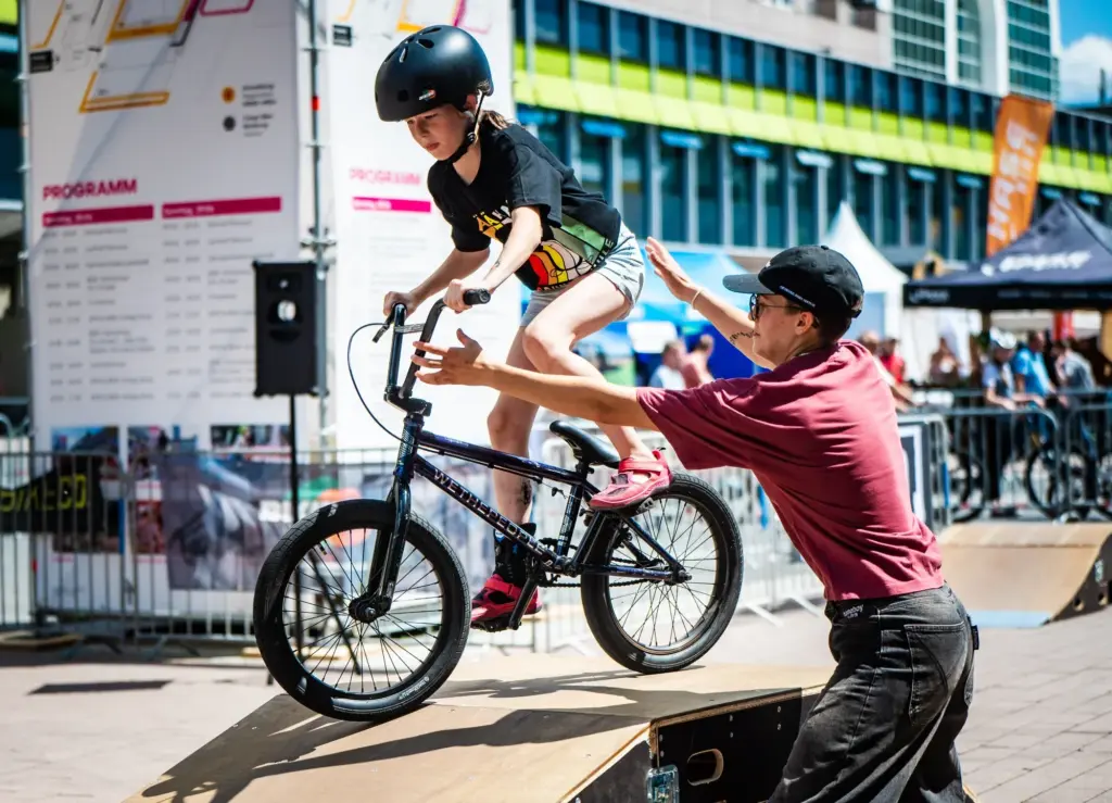 Kind fährt mit BMX-Rad auf einer Holzrampe, ein Erwachsener unterstützt es.