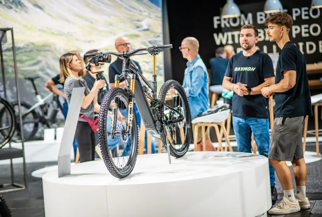 Ein schwarzes E-Bike mit goldfarbener Federgabel steht auf einem runden Podest, flankiert von zwei jungen Männern. Im Hintergrund sind weitere Fahrräder und Besucher einer Messe zu sehen.