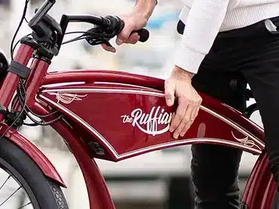 Eine Hand ruht auf dem roten Tank des "the Ruffian" E-Bikes, das als Cruiser mit Retro-Optik beschrieben wird.