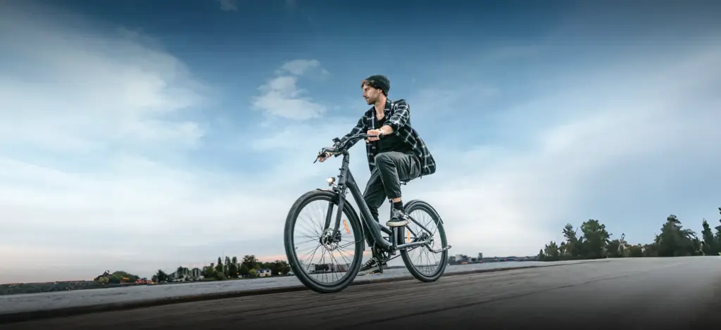 Ein Mann mit Mütze und kariertem Hemd fährt ein schwarzes ADO Air 28 E-Bike auf einem Holzsteg am Wasser.