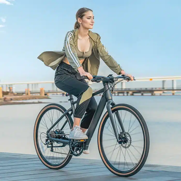 Eine Frau auf einem schwarzen Fiido C21 E-Bike fährt auf einem Steg mit Blick auf eine Brücke und das Meer.