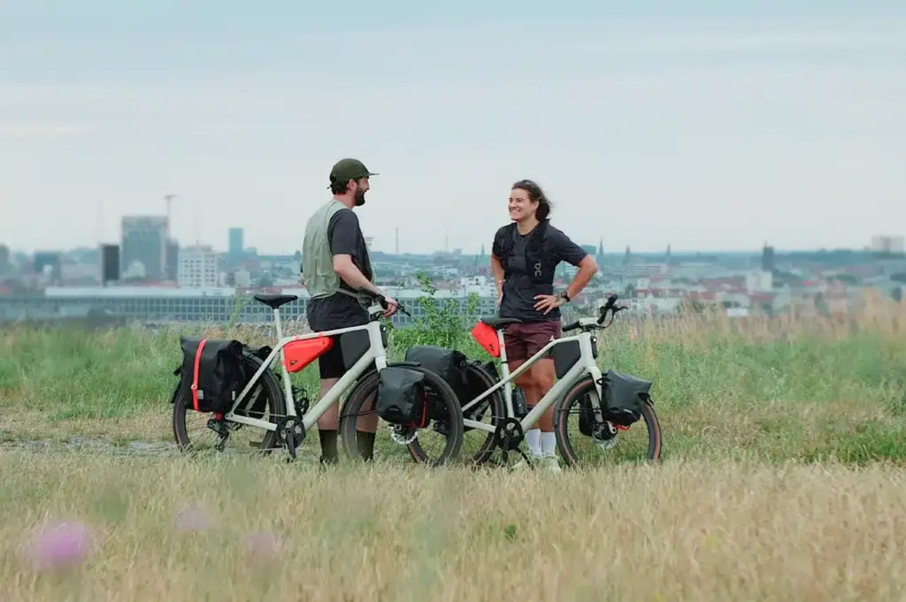 Zwei Personen stehen neben ihren voll ausgestatteten Lemmo One "Collab. Ride" Fahrrädern in einer natürlichen Landschaft mit Stadthintergrund.