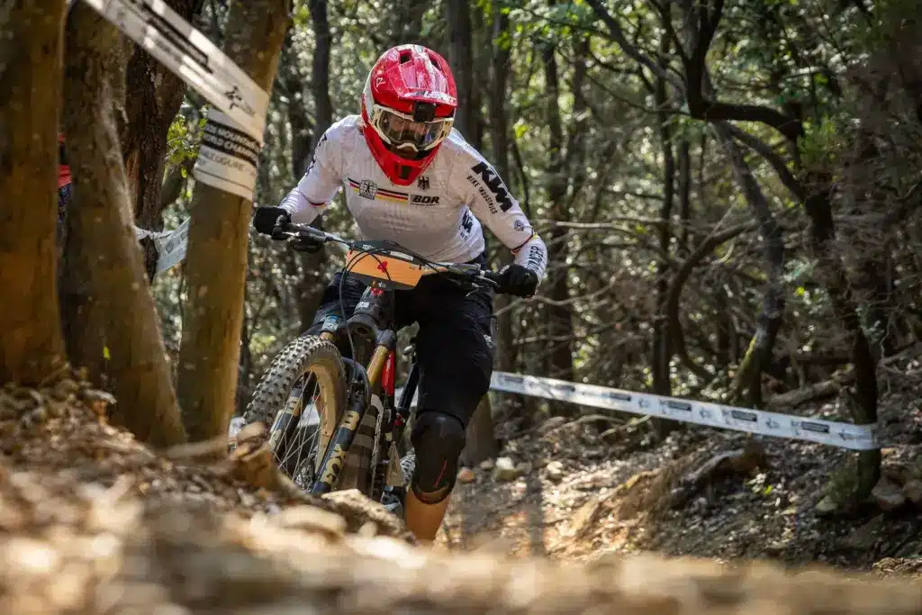 Radsportlerin Helen Weber fährt mit einem roten Helm und einem weißen Trikot auf einem E-Enduro-Mountainbike durch einen Waldweg.