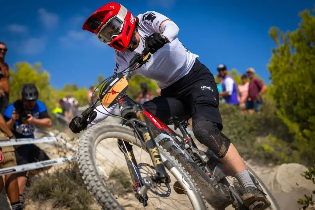 Eine Rennradfahrerin in voller Montur, mit rotem Helm und Schutzbrille, fährt ein E-Enduro-Mountainbike auf einem steinigen Downhill-Kurs. Sie trägt ein weißes Trikot und schwarze Hosen. Im Hintergrund sind Zuschauer und Bäume zu sehen.