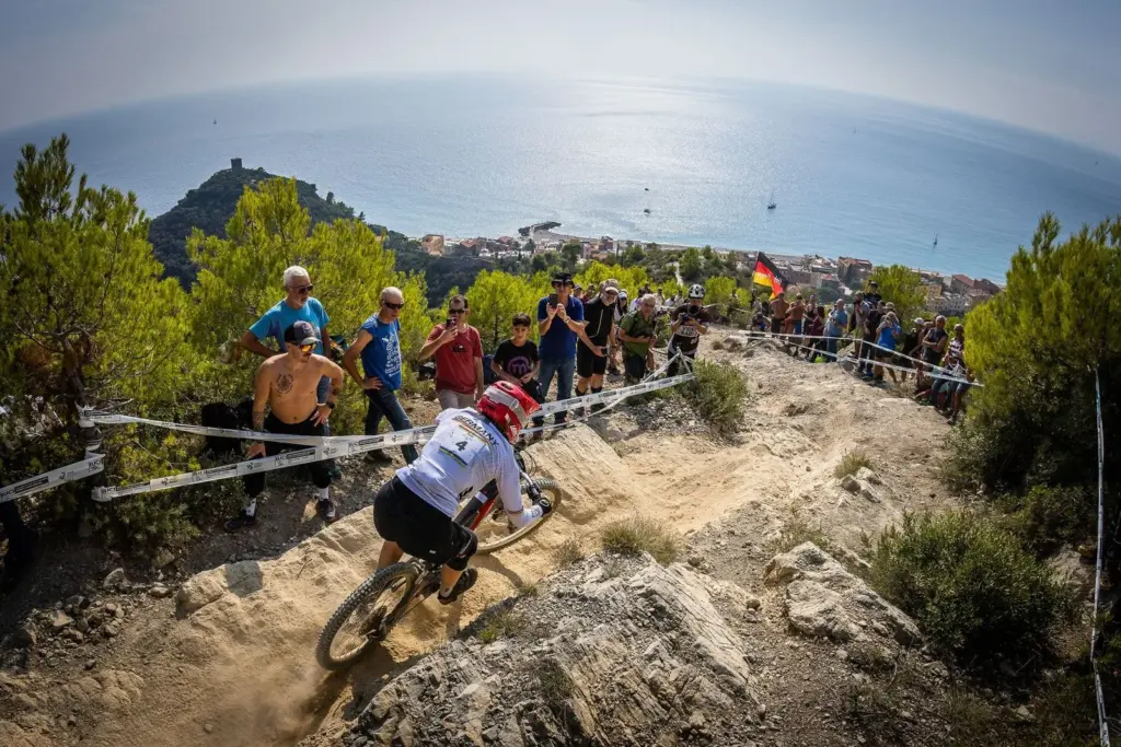 Eine Mountainbikerin mit der Startnummer 4 und der Aufschrift 'Germany' auf dem Trikot fährt einen steinigen Abhang hinunter, während Zuschauer am Streckenrand stehen und das Meer im Hintergrund sichtbar ist.