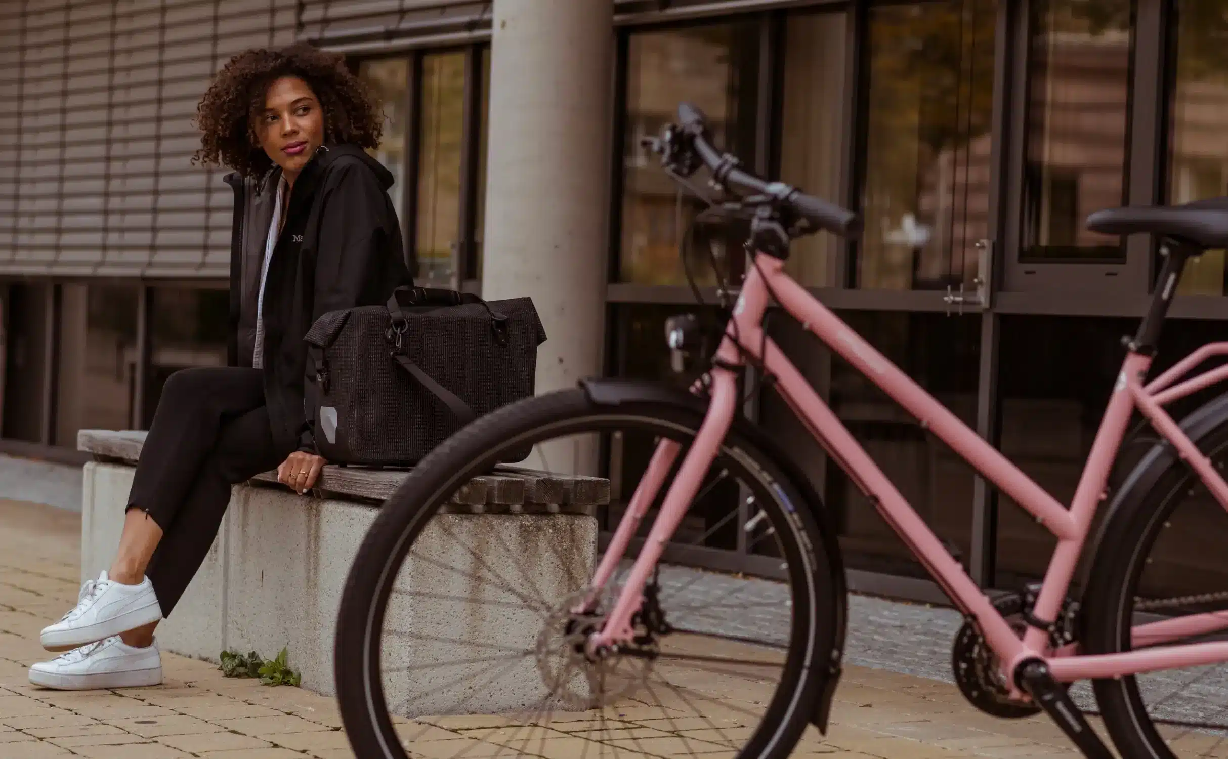 Eine Frau in schwarzer Kleidung sitzt auf einer Betonbank neben einem rosa Fahrrad. Neben ihr steht eine schwarze Fahrradtasche. Das Fahrrad hat eine silberne Felge und schwarze Reifen.