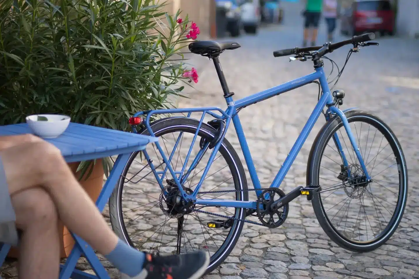 Blaues Trekkingrad der Marke Tout Terrain, Modell Amber Road, mit Gepäckträger und Scheibenbremsen, im Freien in einer Kopfsteinpflasterstraße stehend.