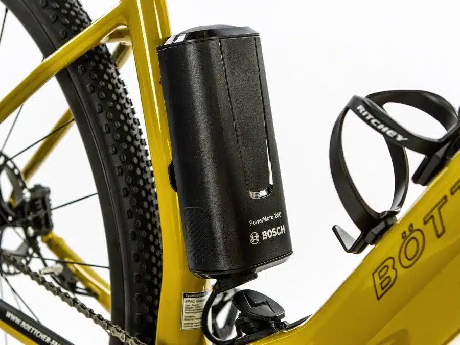 Detailansicht des Böttcher Gravel-E SX Elektrofahrrads mit einem schwarzen Bosch PowerMore 250 Akku, der seitlich am gelben Rahmen befestigt ist. Im Hintergrund ist das Profil des Hinterreifens und ein Teil der Kette zu sehen, sowie eine schwarze Ritchey-Flaschenhalterung.