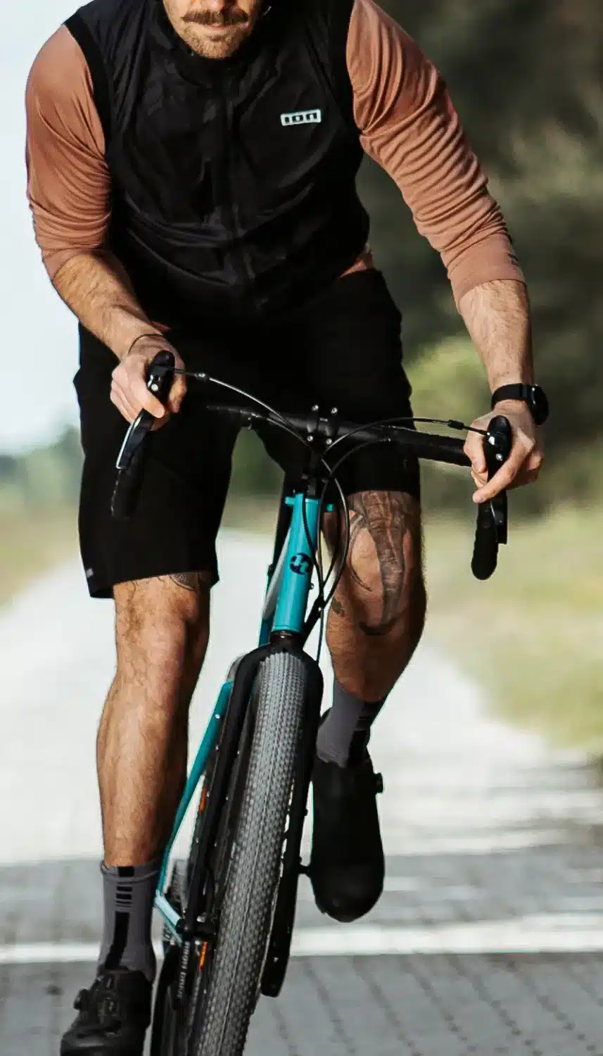 Nahaufnahme eines Mannes mit Bart und Tattoos, der auf einem turkisfarbenen Blueridge Reiserad fährt. Er trägt ein schwarzes Langarmshirt, eine schwarze Weste, schwarze Shorts und Radschuhe. Der Hintergrund ist unscharf und zeigt eine Straße.