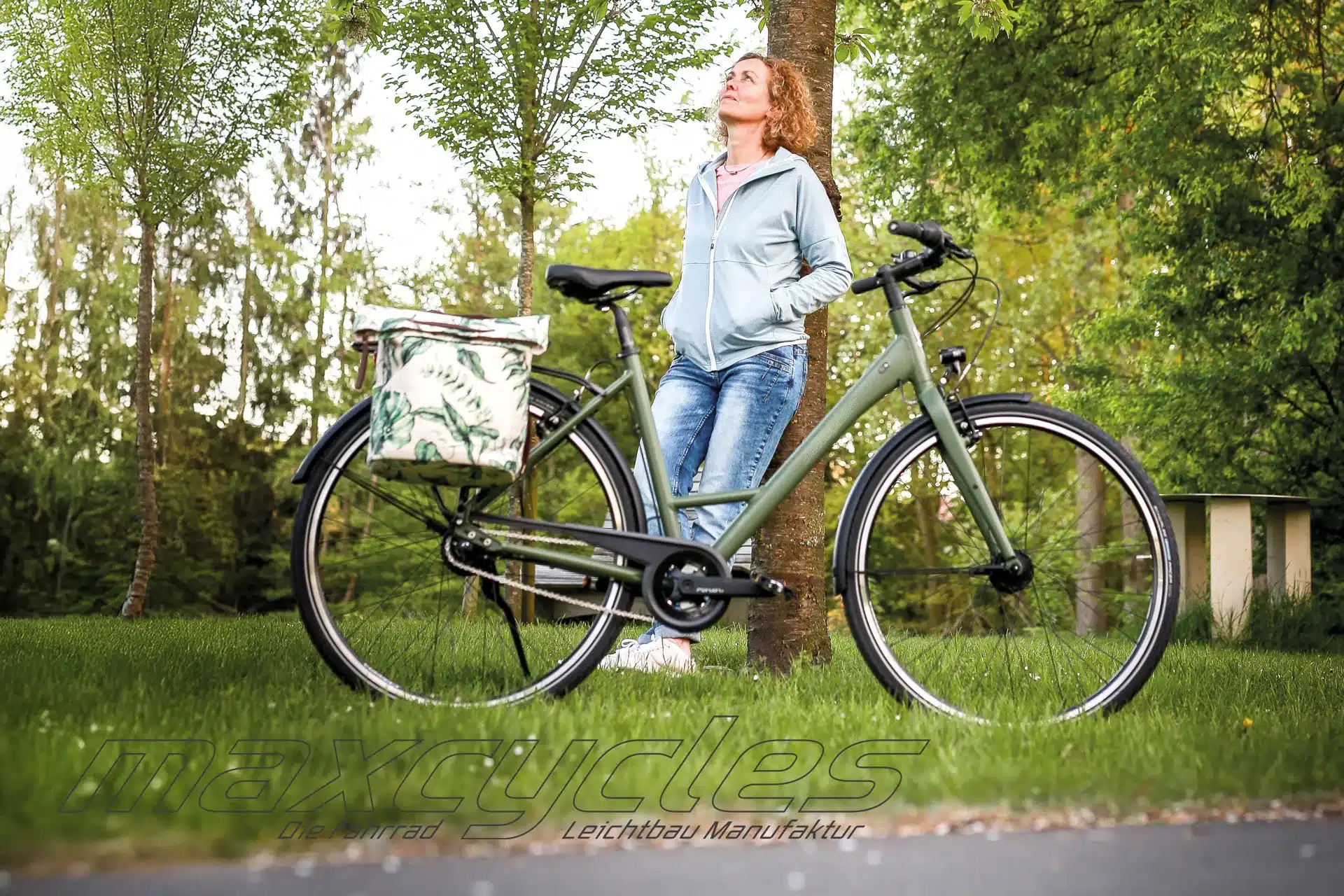 Frau lehnt an Baum neben grünem City-Fahrrad mit Vorder- und Hinterradkorb im Freien