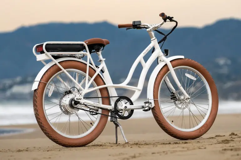 Ein weißes Priority E-Coast Elektrofahrrad mit breiten braunen Reifen und braunem Sattel steht auf Sand am Meer.
