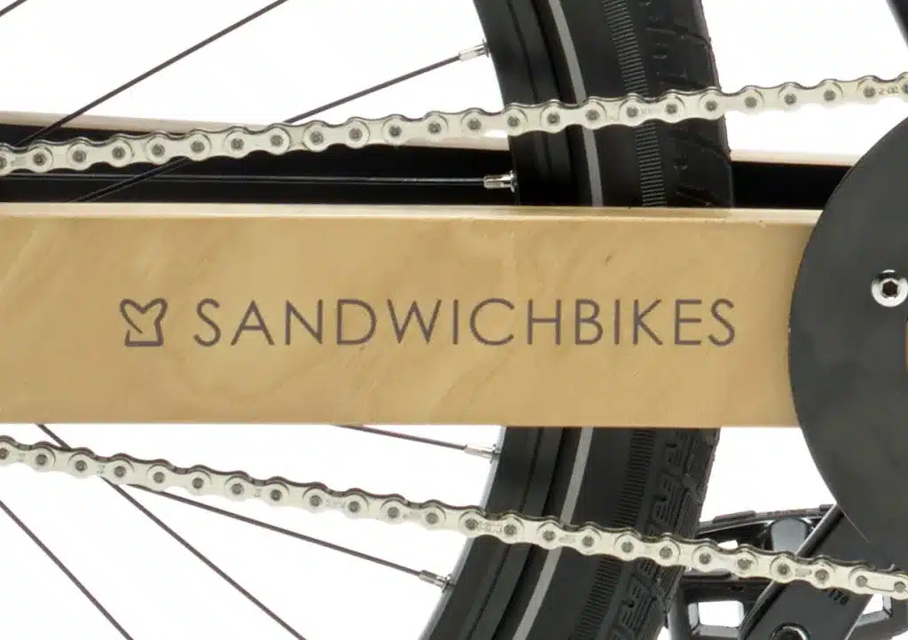 Nahaufnahme des charakteristischen Holzrahmens eines Sandwichbikes, auf dem der Markenname eingraviert ist. Daneben ist die silberne Fahrradkette sichtbar.
