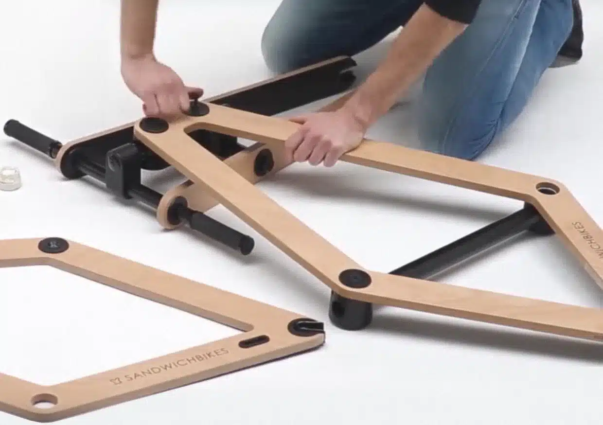 Zwei Hände montieren den Holzrahmen eines Sandwichbikes, wobei die Holzteile mit schwarzen Verbindungsstücken zusammengefügt werden.