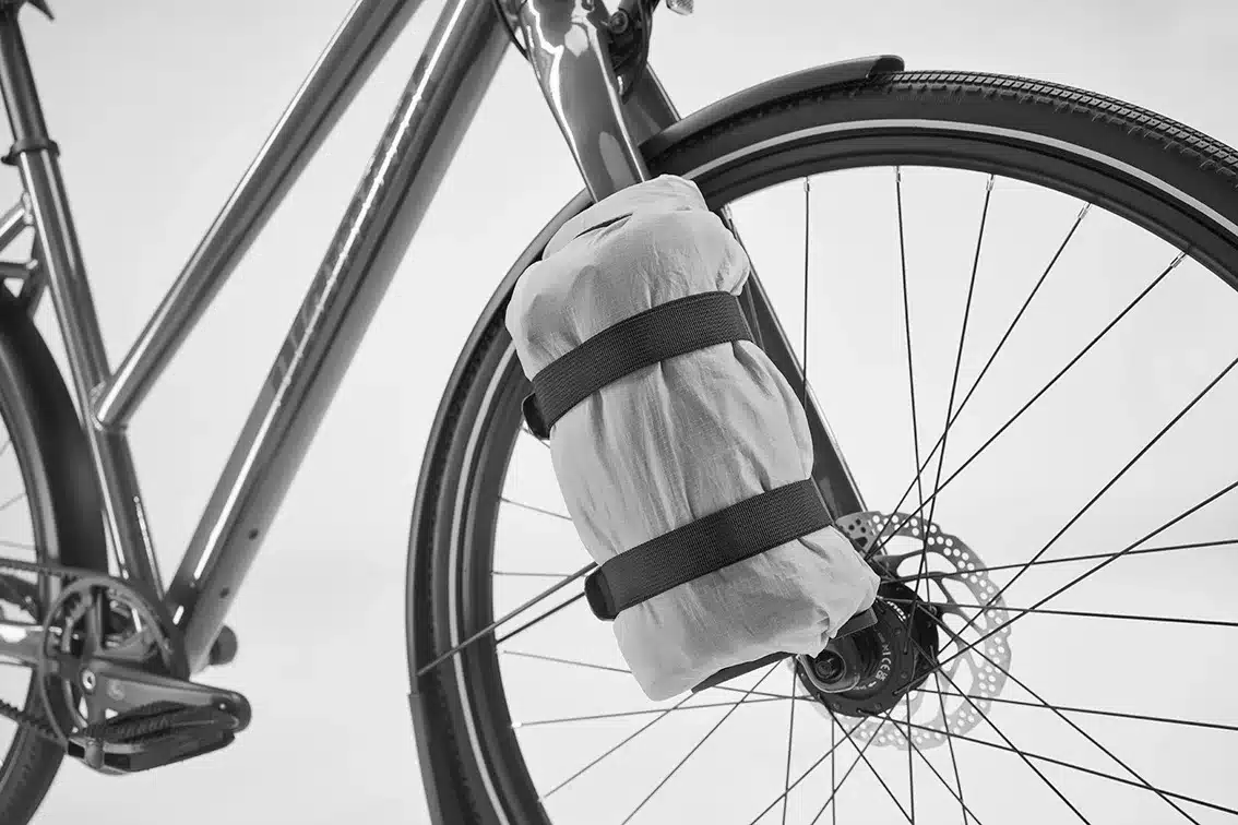 Nahaufnahme des Rahmens eines Fahrrads mit einer zusammengerollten, hellen Tasche, die mit zwei schwarzen Gurten am Steuerrohr und der Vorderradgabel befestigt ist. Der Fokus liegt auf dem Vorderrad, der Scheibenbremse und Teilen des Rahmens.