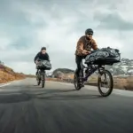 Zwei Gravel-Cargobikes vom Typ Gravit DUST, beladen mit Gepäck, fahren auf einer asphaltierten Straße durch eine Berglandschaft. Die Fahrer tragen Helme.