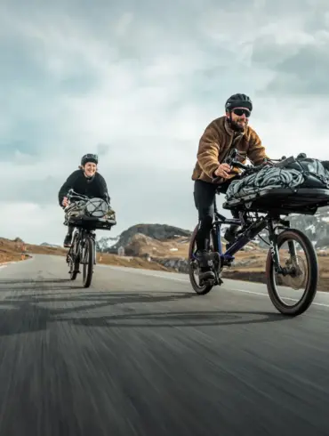 Zwei Gravel-Cargobikes vom Typ Gravit DUST, beladen mit Gepäck, fahren auf einer asphaltierten Straße durch eine Berglandschaft. Die Fahrer tragen Helme.