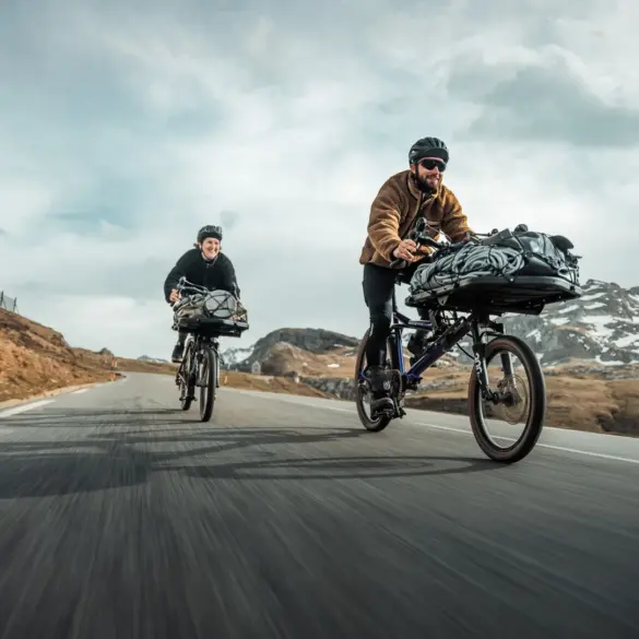 Zwei Gravel-Cargobikes vom Typ Gravit DUST, beladen mit Gepäck, fahren auf einer asphaltierten Straße durch eine Berglandschaft. Die Fahrer tragen Helme.