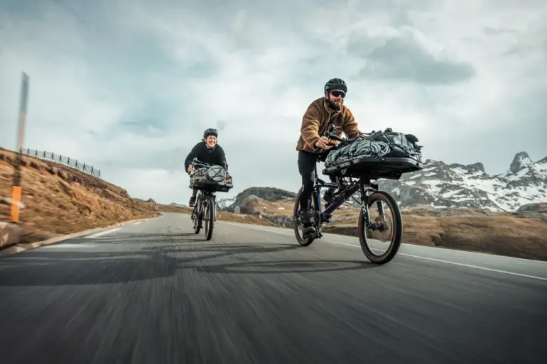 Zwei Gravel-Cargobikes vom Typ Gravit DUST, beladen mit Gepäck, fahren auf einer asphaltierten Straße durch eine Berglandschaft. Die Fahrer tragen Helme.