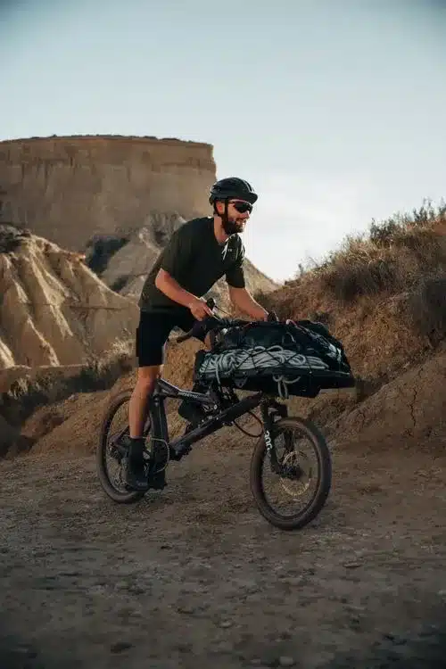 Ein Mann mit Helm und Sonnenbrille fährt ein schwarzes Gravit DUST Lastenrad mit einer Gepäckplattform voller Ausrüstung durch felsiges, trockenes Gelände unter klarem Himmel.