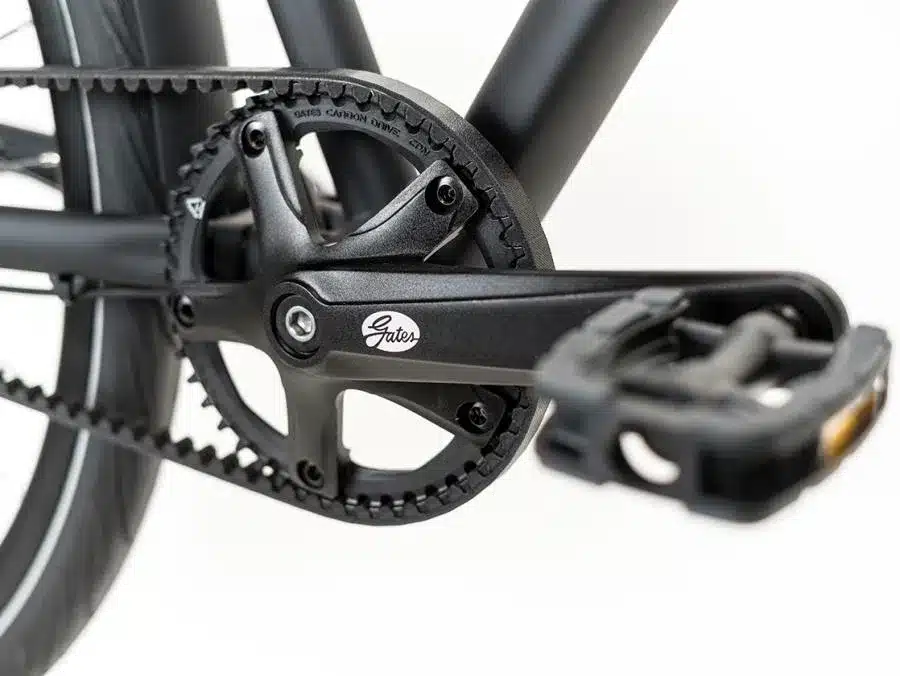 Nahaufnahme des schwarzen Gates Carbon Drive Riemenantriebs und der dazugehörigen Kurbelgarnitur an einem Grecos Urban Belt Diamant Fahrrad, mit Fokus auf den Riemen, das Zahnrad und das Pedal.