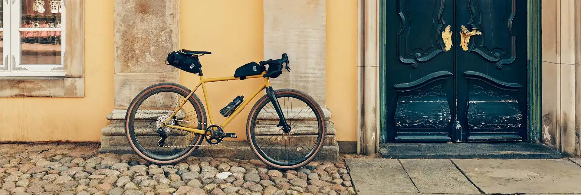 Das GX-1200 1st Edition Gravelbike in Dirty-Mustard-Lackierung mit Bikepacking-Taschen vor einem gelben Gebäude und einer alten Tür
