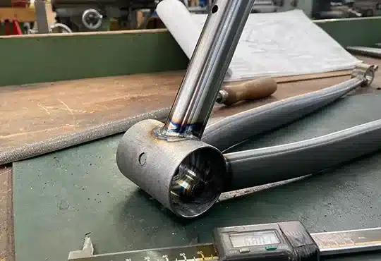 Nahaufnahme eines handgefertigten Stahlrahmens mit sichtbaren Schweißnähten am Tretlager, auf einer Werkbank liegend