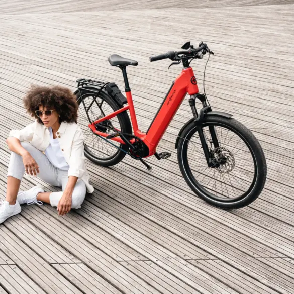 Rotes HNF-NICOLAI UD4 E-Bike mit schwarzem Gepäckträger und Felgenbremsen, neben einer Frau mit Afro-Frisur, Sonnenbrille und weißer Kleidung, die auf einem Holzsteg sitzt.