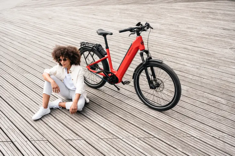 Rotes HNF-NICOLAI UD4 E-Bike mit schwarzem Gepäckträger und Felgenbremsen, neben einer Frau mit Afro-Frisur, Sonnenbrille und weißer Kleidung, die auf einem Holzsteg sitzt.
