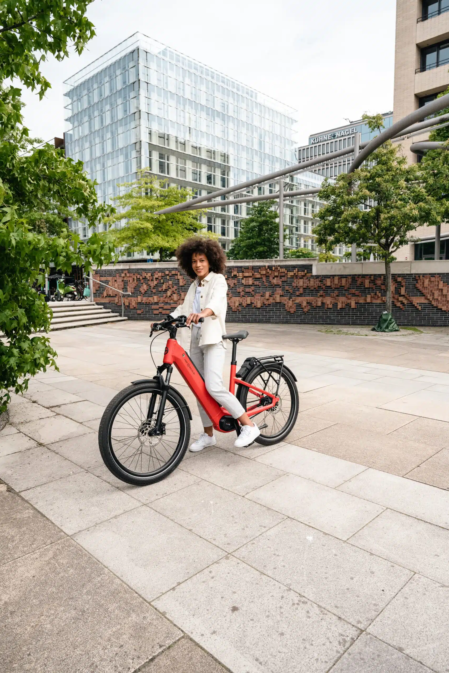 Eine Frau steht mit ihrem roten HNF-Nicolai UD4 E-Bike auf einem Gehweg vor einem großen Glasgebäude.