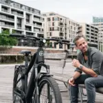 Schwarz lackiertes HNF XD4 E-Bike mit Frontscheinwerfer, Federgabel und integriertem Akku, daneben ein Mann mit grauer Hemd und Jeans auf einer Holzbank im urbanen Umfeld.