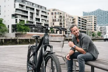 Schwarz lackiertes HNF XD4 E-Bike mit Frontscheinwerfer, Federgabel und integriertem Akku, daneben ein Mann mit grauer Hemd und Jeans auf einer Holzbank im urbanen Umfeld.