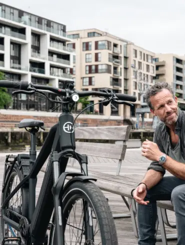 Schwarz lackiertes HNF XD4 E-Bike mit Frontscheinwerfer, Federgabel und integriertem Akku, daneben ein Mann mit grauer Hemd und Jeans auf einer Holzbank im urbanen Umfeld.