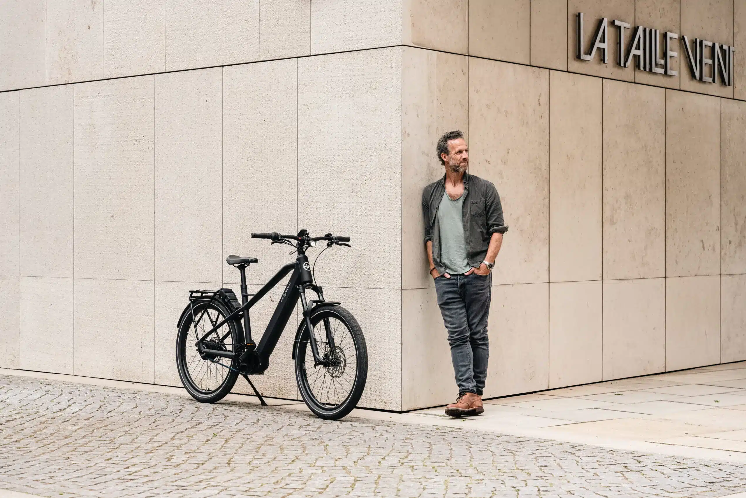 Schwarzes HNF XD4 E-Bike mit Gepäckträger steht neben einem stehenden Mann im Freien vor einer modernen Gebäudewand aus hellen Natursteinplatten.